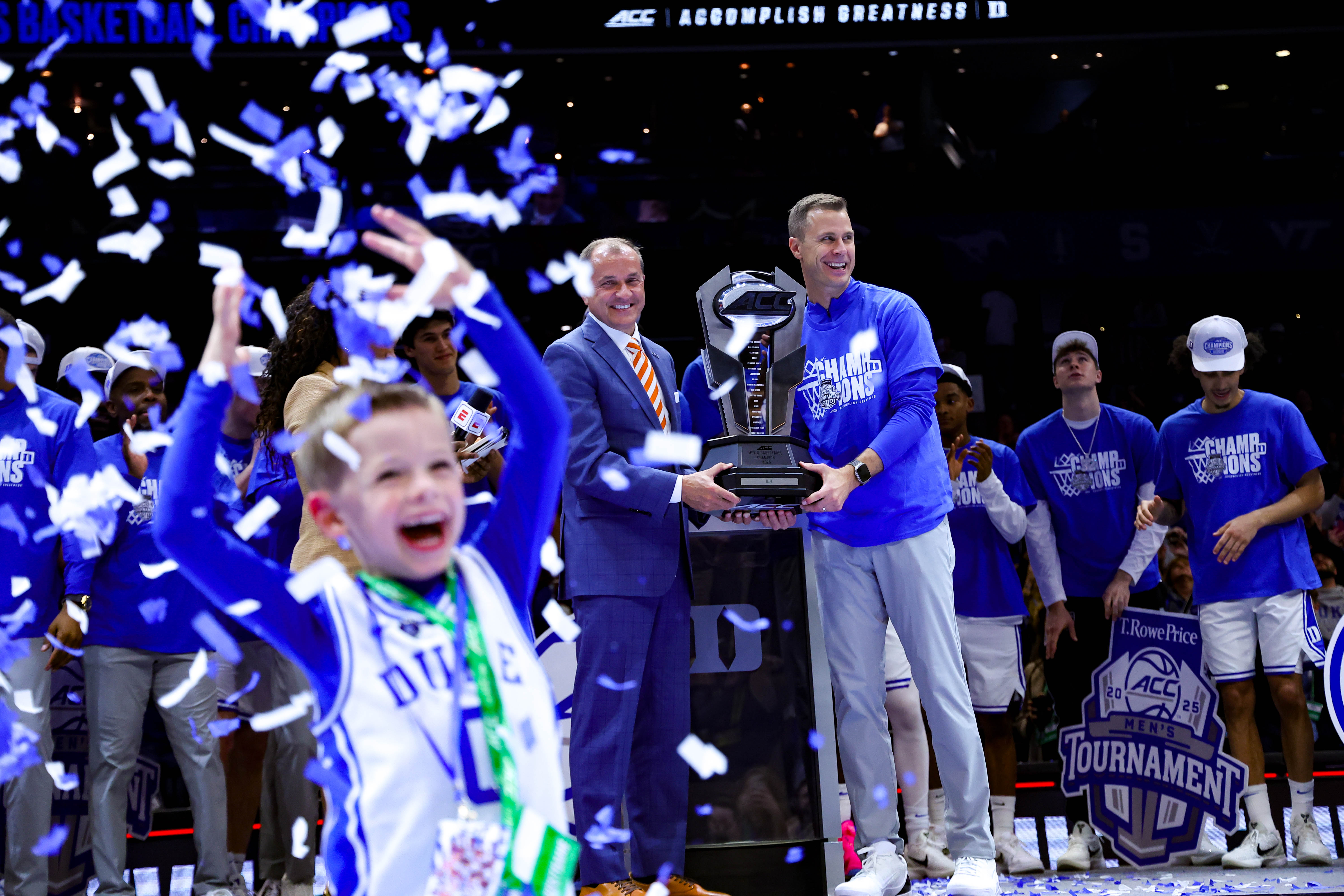 Jon Scheyer and Jim Phillips pose with the ACC Basketball Tournament Championship trophy as fans celebrate Duke winning the conference tournament. (Photo credit: ACC Communications)